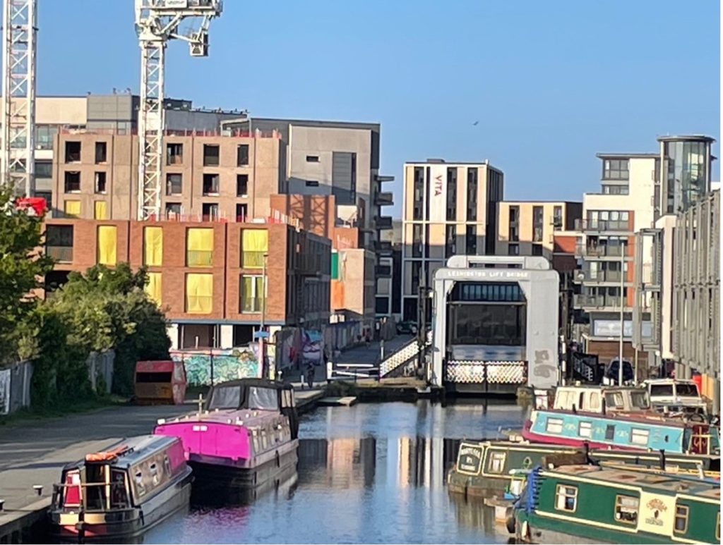 A photograph of Leamington Lift Bridge and the nearby canal-side. The skies are blue and there are several canal boats in view.